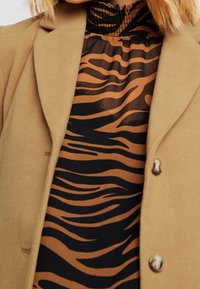 Tan uldblandingsblazer med slået krave og metalliske knapper, lagd ovenpå en sort og brun stribet bluse med høj hals.