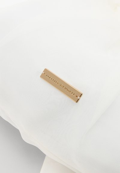 Bijela tkanina s glatkom teksturom, koja sadrži pravokutnu zlatnu metalnu oznaku s reljefnim natpisom "LOEFFLER RANDALL."