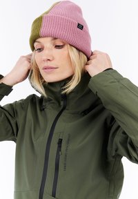 Vrouw die de kraag van een olijfgroene waterdichte jas aanpast, met een roze en olijfkleurige gebreide muts met een "House of Snow" label.