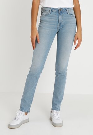 Lichtblauwe slim-fit jeans gedragen met witte sneakers en een wit shirt, getoond van taille tot voeten tegen een effen achtergrond.