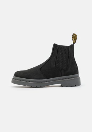 Zwarte suède Chelsea boot met elastische zijpanelen en een trekband. Dikke grijze rubberen zool met een gestructureerd profiel voor grip. Minimalistisch ontwerp.