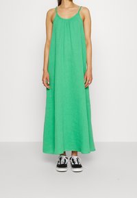 Zielona, bezrękawnikowa sukienka maxi wykonana z teksturowanej tkaniny, o luźnym kroju. Połączona z czarno-białymi sneakersami.
