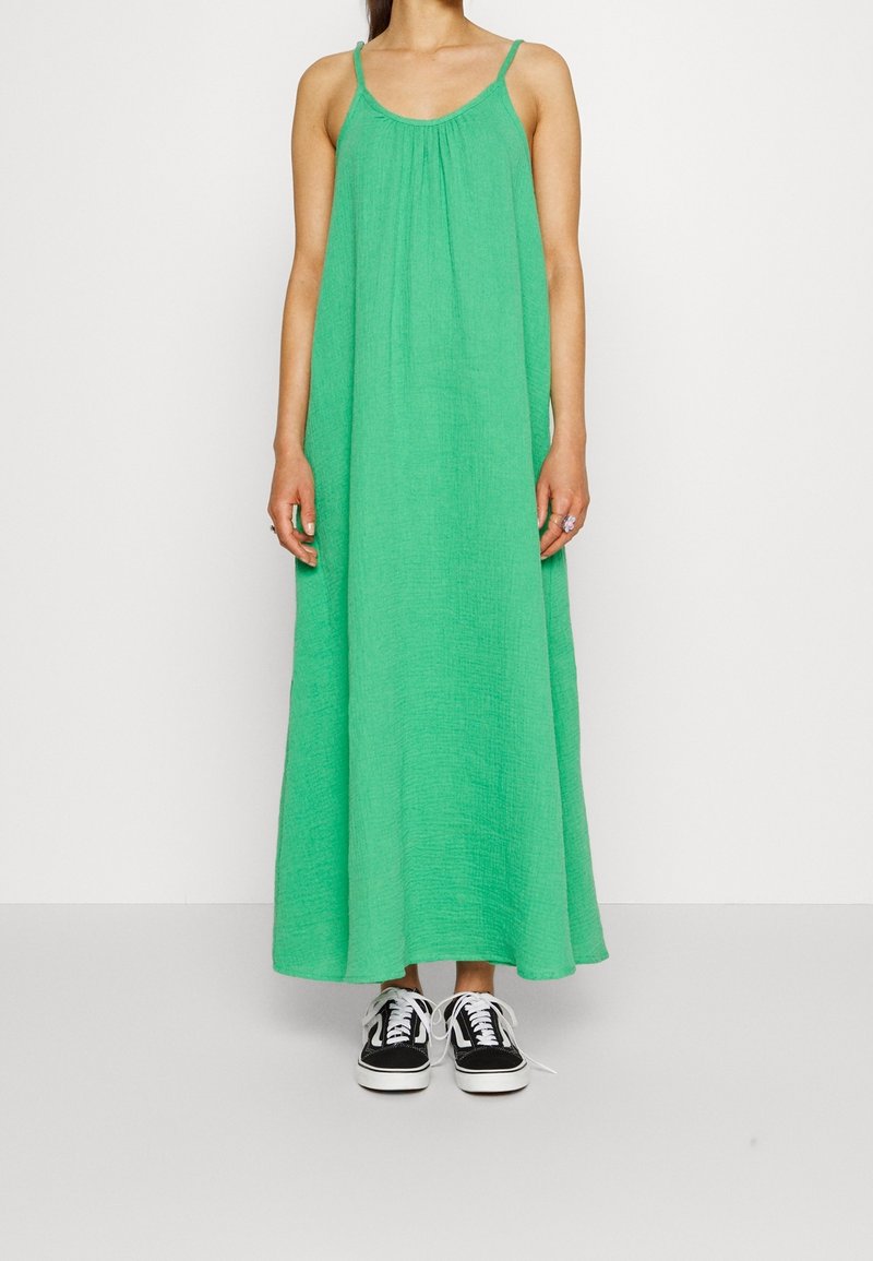 Zielona, bezrękawnikowa sukienka maxi wykonana z teksturowanej tkaniny, o luźnym kroju. Połączona z czarno-białymi sneakersami.