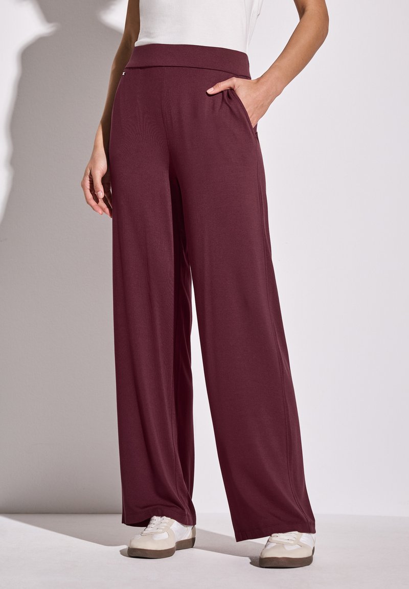Pantalons larges bordeaux avec une texture douce, taille élastique et poches latérales, assortis à des baskets blanches sur une surface claire.