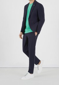 Mørkeblå blazer, grøn t-shirt, mørkeblå bukser og hvide sneakers. Blazeren har en enkelt knap og en brystlomme; glat stof generelt.