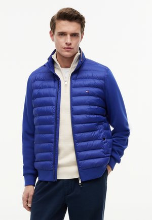 Ung mand iført en blå quiltet jakke over en beige halv lynlås sweater og mørke bukser, stående med den ene hånd i lommen.