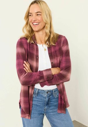 Smiling woman with blonde hair wears a purple plaid shirt over a white top and blue jeans, standing with arms crossed against light background.