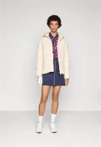 Moteris su trumpais garbanotais plaukais vilki kreminio atspalvio pūkinę striukę, spalvingą polo marškinėlį, tamsiai mėlyną sijoną su baltu užtrauktuku, baltas kojines, sportinius batelius ir pirštinę.