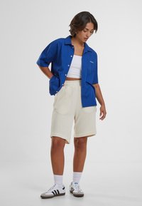 Blå kortærmet skjorte med knapper og lomme, cremefarvede strikkede shorts og lave sneakers. Hvide sokker synlige, afslappet outfit med løs pasform.