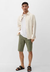 Jasno kremowa, lekka kurtka na guziki nałożona na białą koszulkę, w połączeniu z oliwkowymi szortami i czarnymi sandałami. Prosty, casualowy design.