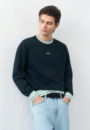 Młody mężczyzna ubrany w czarną bluzę z miętowozielonymi mankietami i kołnierzem, jasnoniebieskie dżinsy, ręce w kieszeniach, patrzący w dół w lewą stronę.