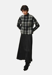 Osoba ubrana w sweter w czarno-białą kratę, długą czarną spódnicę i czarne botki za kostkę stoi z rękami za plecami na tle białego tła.