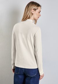 Långärmad krämfärgad topp med hög krage, gjord av ett mjukt tyg. Har en avslappnad passform, kombinerad med mörkblå jeans.