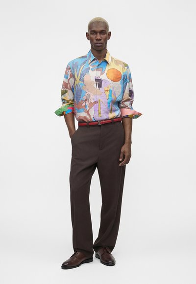 Άνδρας με κοντά ξανθά μαλλιά που φοράει πολύχρωμο πουκάμισο με αφηρημένο μοτίβο, καφέ παντελόνια, κόκκινη ζώνη και σκούρα καφέ επίσημα παπούτσια.