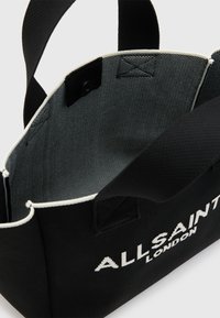 Borsa nera con il testo bianco "ALLSAINTS LONDON"; tessuto morbido, forma strutturata, manici corti e un interno grigio con un motivo tessile.