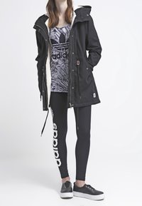 Black parka jacket with a white fleece lining, paired with a grey and black graphic tank top, black leggings with white logo, and black sneakers.