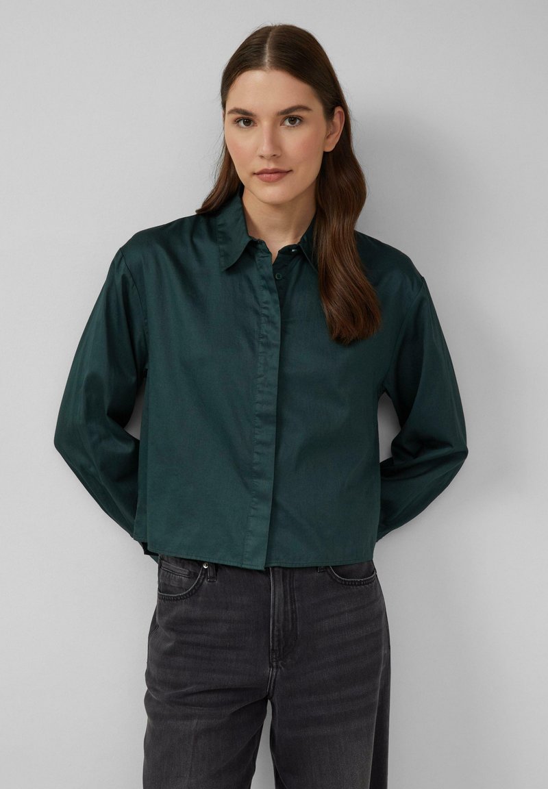 Chemise courte vert foncé en tissu lisse, dotée d'un devant à boutons, d'un col classique et de manches longues, accompagnée d'un jeans gris foncé.