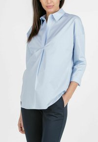 Helles blaues Button-Up-Hemd aus Baumwollmischung, das ein geteiltes Design an der Vorderseite und hochgekrempelte Ärmel aufweist, kombiniert mit dunklen Hosen.