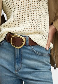 Bruine leren riem met een grote, ovale gouden gesp. Gecombineerd met een lichte gebreide trui en blauwe denim jeans.