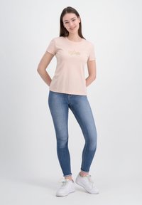 Růžové tričko s krátkým rukávem s grafickým designem zlaté hvězdy a textem, kombinované s modrými skinny džíny a bílými teniskami.