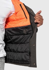Chaqueta aislante con un exterior gris, interior naranja y sección inferior negra. Incluye bolsillos con cremallera y una superficie texturizada.