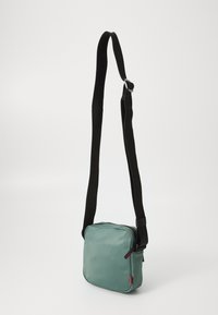 Borsa a tracolla quadrata verde con una tracolla nera, chiusura con zip e un piccolo dettaglio rosso. Realizzata in materiale liscio e resistente.
