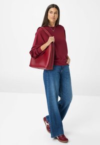 Bordeaux sweatshirt gecombineerd met wijdvallende blauwe jeans en bordeaux sneakers, terwijl een rode draagtas wordt vastgehouden, tegen een effen witte achtergrond.