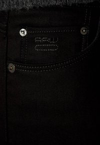 Mustat farkut, joissa etutasku hopeisella metalliosalla ja ompeludetaljeilla. Kangas on sileä ja tukeva.