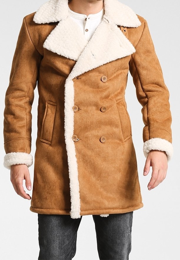 Homme portant un manteau croisé beige avec col, poignets et bordure en shearling blanc, associé à un pantalon gris foncé.