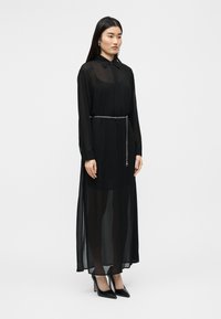 Μακρύ μαύρο διάφανο φόρεμα με γιακά και κουμπωτή πατιλέτα, εφαρμοστό στη μέση με ασημένια αλυσίδα-ζώνη. Το μοντέλο φοράει μαύρες ψηλοτάκουνες γόβες.