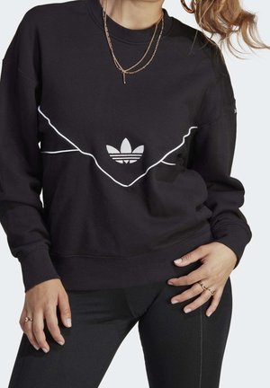 Kobieta w czarnej bluzie Adidas z białym logo i białymi paskami, czarne spodnie, złoty warstwowy naszyjnik i pierścionki na obu rękach.