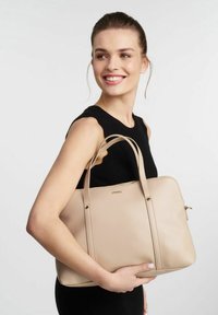 Beżowa skórzana torba typu tote z złotymi akcentami, o uporządkowanej konstrukcji, z podwójnymi uchwytami i gładką teksturą. Zamek błyskawiczny oraz minimalne detale.