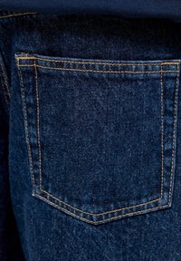 Poche de jean en denim bleu foncé avec surpiqûres orange visibles à l'arrière du vêtement, montrant des détails texturés du tissu.