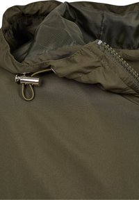 Capuche de veste verte avec bouton-pression métallique argenté et cordon près d'une fermeture éclair partiellement visible sur un matériau en tissu.