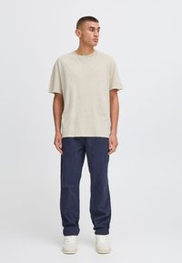 Beige knit t-shirt with short sleeves, paired with navy blue pants and white sneakers. Model stands upright on a plain background.