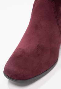 Gros plan d'une botte cheville en daim bordeaux avec un bout arrondi et une semelle noire sur fond blanc.
