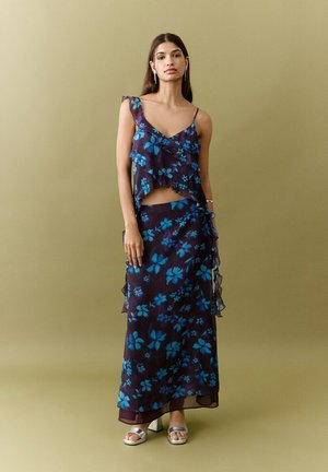 Femme portant une tenue deux pièces fleurie violet foncé avec des volants, debout devant un fond beige uni, avec des talons argentés ouverts à l'avant.