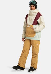 Kurtka snowboardowa z wielokolorowymi panelami (kremowym, burgundowym, miętowym) i kieszenią z przodu. W połączeniu z beżowymi spodniami cargo i czarnymi butami snowboardowymi.