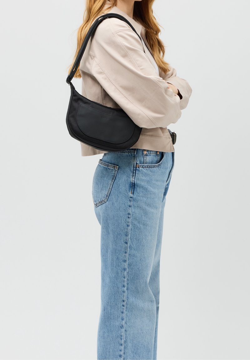 Svart axelväska med en böjd form och en slät yta. Har en lång rem och en liten framficka. Bärs med en ljus outfit.