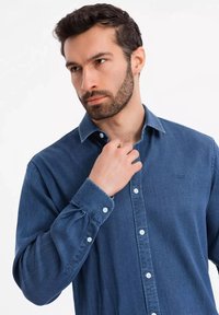 Camicia di jeans in tessuto blu scuro, maniche lunghe, colletto abbottonato e bottoni bianchi; presenta un piccolo logo nella zona della tasca sul petto.