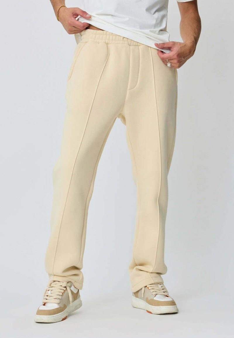 Pantalons de jogging beiges en tissu doux, dotés de poches latérales et d'une coupe décontractée. Portés avec un t-shirt blanc et des baskets aux tons neutres.