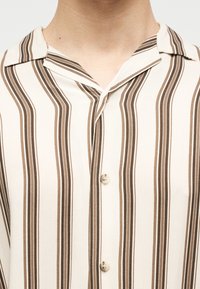 Πουκάμισο με γιακά, με κάθετες καφέ και μπεζ ρίγες, κουμπωμένο μπροστά, ορατό από το λαιμό μέχρι το μέσο του στήθους.