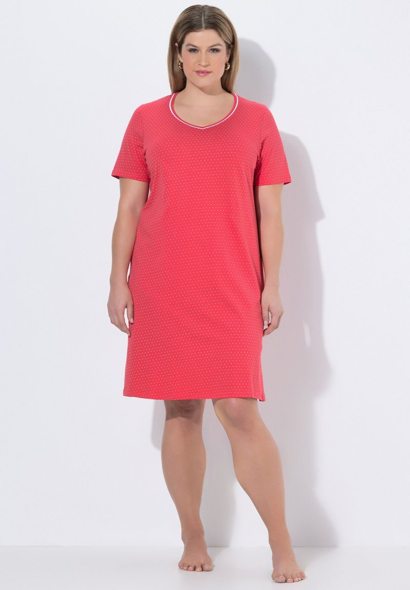Femme debout pieds nus portant une robe rouge mi-longue à manches courtes avec de petits pois blancs et un col bordé de blanc.