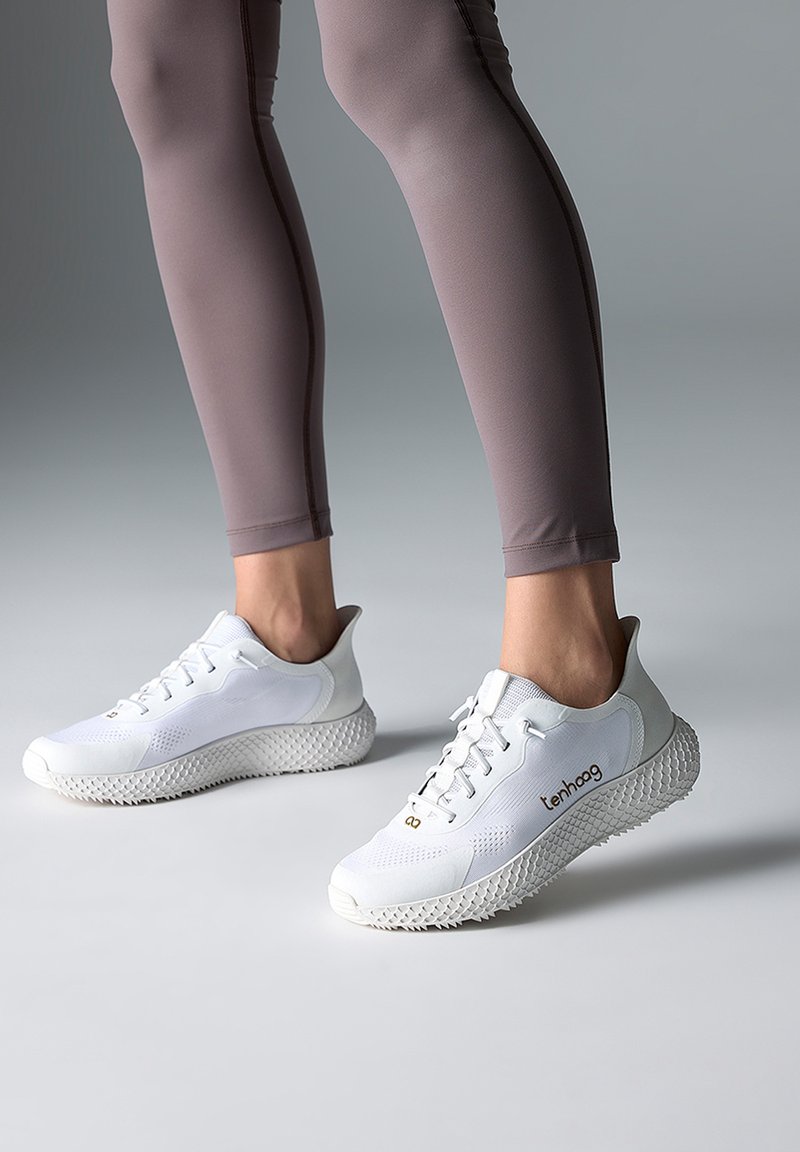 Ben i grå leggings iført hvide sportssko med teksturerede såler og "tenhoag"-mærke, stående på en glat grå overflade.