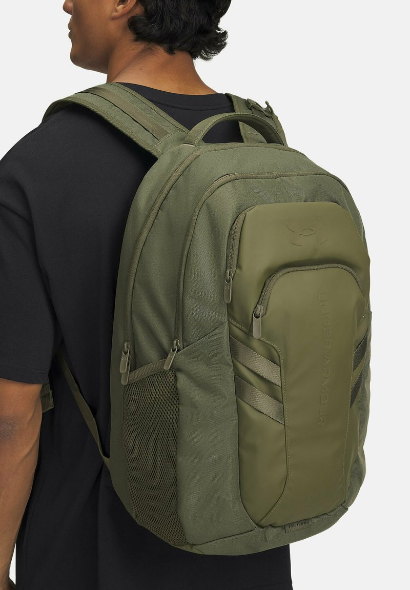 Olivgrüner Rucksack mit mehreren Fächern, einer seitlichen Netztasche und gepolsterten Trägern. Verfügt über das "Under Armour"-Logo und strukturierte Oberflächen.