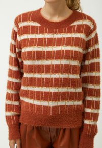 Gestrickter Pullover in Rost mit weißen und goldenen Streifen. Weiche, flauschige Textur mit einem gerippten Kragen und Bündchen. Boxy-Schnitt, mittellang.