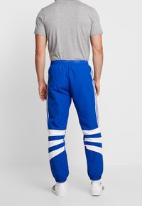 Blå sportbyxor med vita diagonala ränder, elastisk midja och ribbade muddkanter. Bärs med en grå t-shirt med kort ärm och vita skor.