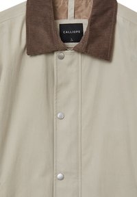 Giacca beige con colletto in velluto a coste marrone, chiusure a pressione e una texture di tessuto liscia. Etichetta che indica "CALLIOPE," taglia L.