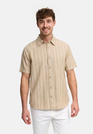 Uśmiechnięty mężczyzna w beżowej koszuli z krótkim rękawem z białymi pionowymi prążkami oraz białych spodniach, stojący na tle jednolitego jasnego tła.