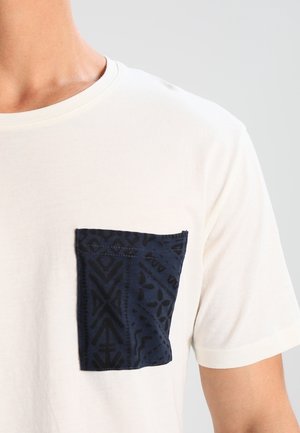 Mand iført en hvid t-shirt med en mørkeblå mønstret brystlomme på venstre side, foran en ensfarvet lys baggrund.